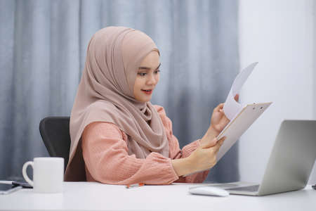Happy asian muslim woman brown hijab working at home with laptop and documents.の写真素材