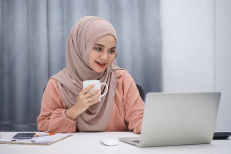 Beautiful asian muslim woman brown hijab drinking coffee and making a phone call while working at her home.の写真素材