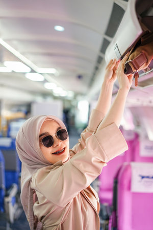 Beautiful muslim female traveler passenger putting her hand luggage on overhead locker before takeoff.の写真素材