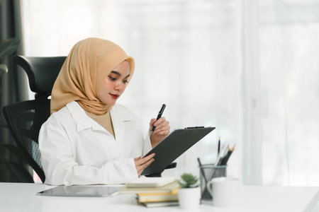 A smiling and confident young Muslim businesswoman auditor is writing on a clipboard and signing a contract document at her desk in a modern office.の写真素材
