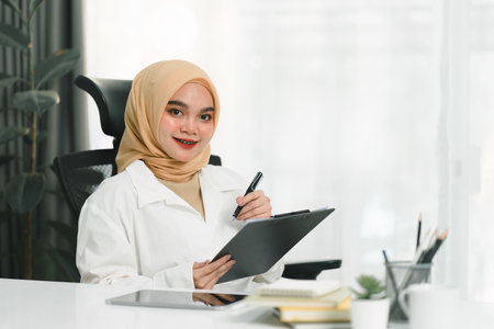 A smiling and confident young Muslim businesswoman auditor is writing on a clipboard and signing a contract document at her desk in a modern office.の写真素材