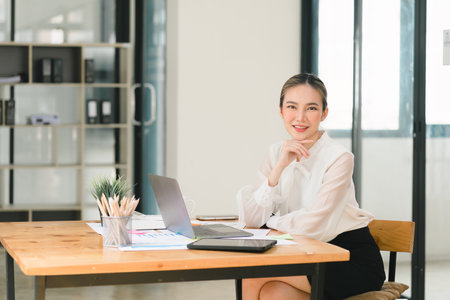 An attractive young businesswoman is using a laptop in a modern office and looking at the camera.の写真素材