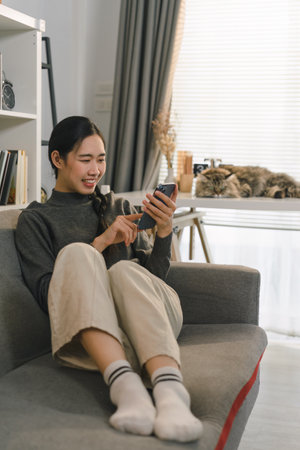 Charming Asian woman is using her smartphone while relaxing on the sofa at home.の写真素材