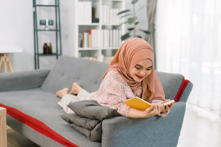 A portrait of a smiling Muslim woman wearing a hijab while reading a book on a couch in the living room of her home.の写真素材