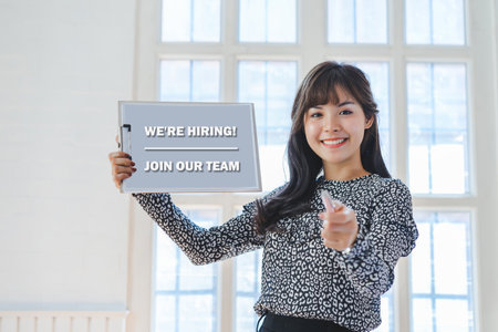 Happy young asian woman hand holding paper with text WE ARE HIRING,JOIN OUR TEAM and finger pointing towards the camera.の写真素材