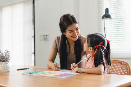 Young mother assists her daughter with coloring activities at a home desk during daytime