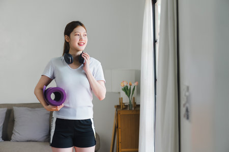 A cheerful woman stands in the living room, holding a purple yoga mat and wearing headphones, ready for a yoga session.の写真素材