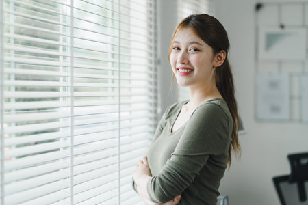 Smiling asian young businesswoman standing by the window in a bright home officeの写真素材