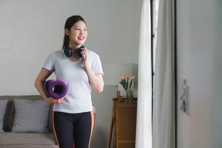 A cheerful woman stands in the living room, holding a purple yoga mat and wearing headphones, ready for a yoga session.の写真素材
