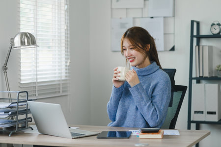 Smiling young woman enjoying a warm beverage during a break in a modern home officeの写真素材