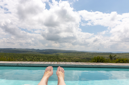 A Close up view of a women's feet with a magical view of a crystal blue pool and vast african bush leading into the mountainsの写真素材