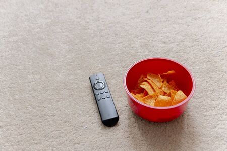 a close up view of a smart tv remote next to a red plastic bowl of chips or crisps on the light coloured carpetの写真素材
