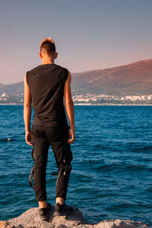 Handsome young man in a black T-shirt and shorts stands on the edge of a cliff and looks at the sea