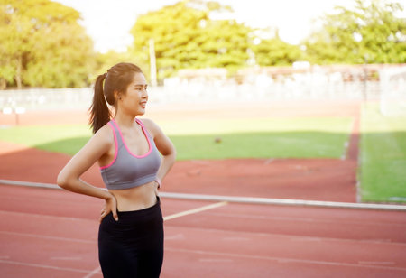 Young Asian female relaxing after workout and jogging at the football stadium in the morning with sun flare background.の写真素材