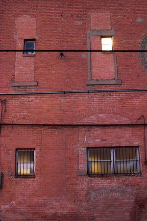Four different windows on a grungy red brick wall in an alley.  One upper window has a light brightly shining which contrasts with the dim glow from the other windows.  Lower two windows have security bars.の写真素材