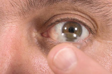 Closeup view of a man's brown eye while inserting a corrective contact lens on a finger with a white background.  Focus is on the eye and the finger and contact lens are out of focus.の写真素材