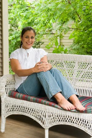 Beautiful smiling young brunette woman with her hair pulled back sitting barefooted comfortably on a love seat on the porch.の写真素材
