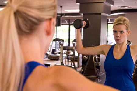 Beautiful blond woman lifting weights while looking in a mirror in a fitness center. Reflection in mirror is in focus - foreground image is out of focus.の写真素材