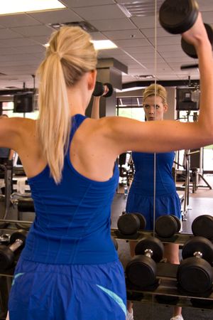 Beautiful blond woman lifting weights while looking in a mirror in a fitness center. Reflection in mirror is in focus - foreground image is out of focus.の写真素材