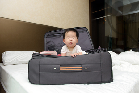 Asian baby sitting in travel suitcase placed on hotel double bedの写真素材