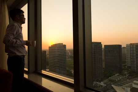 Thoughtful businessman holding a cup of coffee standing by the glass windowの写真素材