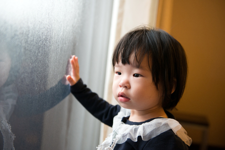 Adorable Asian toddler watching the snow through windowの写真素材