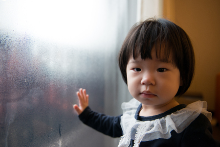 Adorable little unhappy kid standing by windowの写真素材