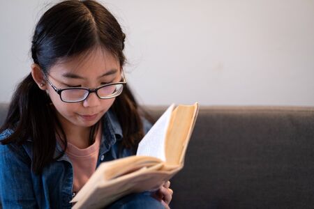 Asian teenage girl reading book and sitting on couch at homeの写真素材