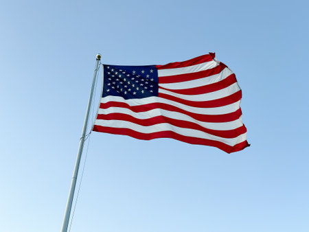 US American flag waving in the wind with a blue sky in background, view from ground level.
