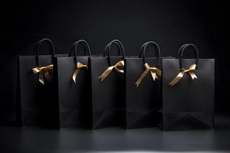 Multiple black shopping and gift bags tied with gold ribbons isolated on dark backgroundの素材