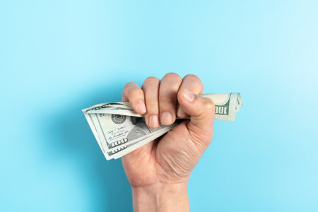 Top view a hand in a fist squeezing cash money hundred dollar bills isolated blue background, concept of no enough money or inflationの写真素材