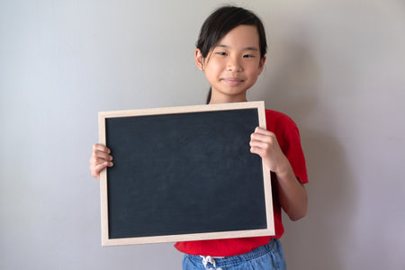 Happy Asian Chinese little girl smiling and hold blank blackboard isolated on grey background. Blackboard for advertising text, copy space.の写真素材