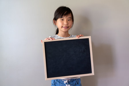 Happy Asian Chinese little girl smiling and hold blank blackboard isolated on grey background. Blackboard for advertising text, copy space.の写真素材