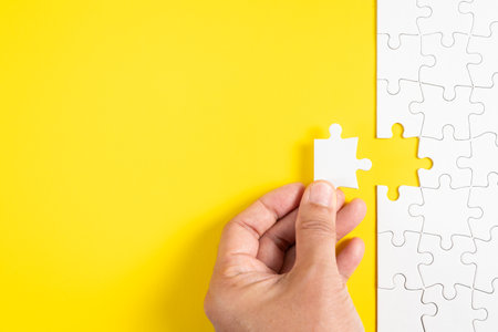 A hand placing the final piece of a white puzzle against a bright yellow background. It symbolizes the completion of a task, problem solving, and the satisfaction of fitting the last piece into place.の写真素材