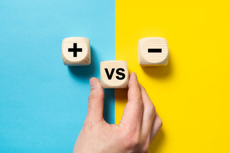 Hand placing a wood block versus between plus and minus sign on blue and yellow background, concept of opposites, pros and cons, lay flat compositionの写真素材