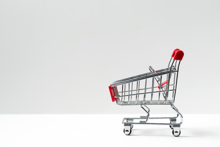 Side view closeup of shiny aluminum empty miniature shopping supermarket cart with red handle isolated on white backgroundの写真素材