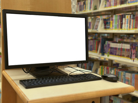 A public computer station in a library with a blank monitor screen, keyboard, and mouse on a wooden desk, surrounded by bookshelves filled with books.の写真素材