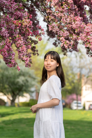 A portrait of a girl teen in a white dress smiling under a tree covered in vibrant pink flowers, set in springtime.の写真素材