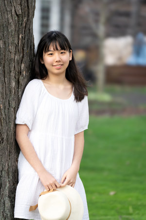 A portrait of a girl teen with hat in a white dress lean against tree, set in springtime.の写真素材