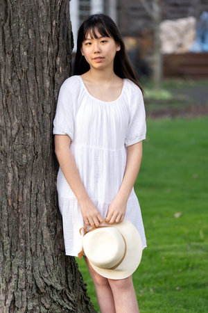 A portrait of a girl teen with hat in a white dress lean against tree, set in springtime.の写真素材