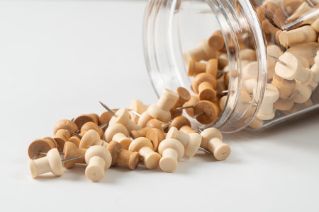 A clear glass jar is tipped on its side, with a collection of wooden pushpins spilling out onto a white surface. The pushpins are a mix of light and medium wood tones, each with a silver metal point.の写真素材