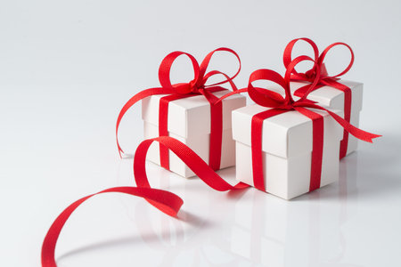 Three square white gift boxes tied with a red satin ribbon for Christmas, New Year, valentines or birthday theme. The ribbon trails around the box on a reflective white surfaceの写真素材