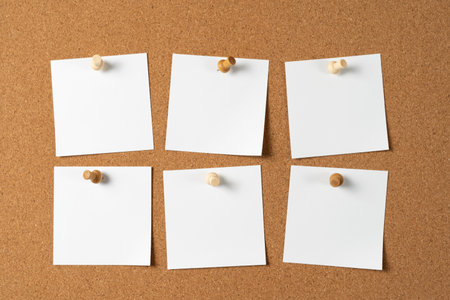 Six blank white notes pinned to a cork bulletin board with wooden push pins, creating space for reminders, messages, or ideas in an organized layout.の写真素材