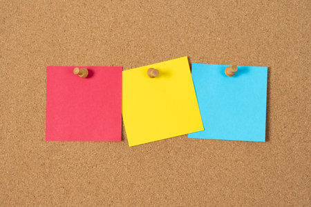 Three blank sticky notes in red, yellow, and blue are pinned to a cork board, ready for messages or reminders.の写真素材