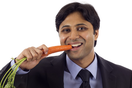 Young Indian business man eating an organic carrot reward, isolated over white backgroundの写真素材