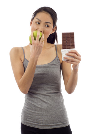 Healthy Choice:  Young woman eating green apple and looking at the chocolate bar isolated over white backgroundの写真素材
