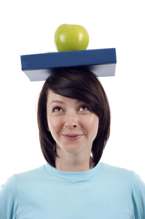 Happy friendly female student looking up with a blue book and green apple on her head isolated whiteの写真素材