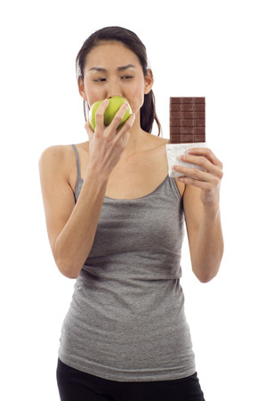 Healthy Choice:  Young woman eating green apple and looking at the chocolate bar isolated over white backgroundの写真素材