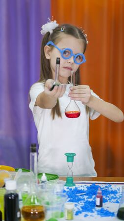 Child is holding flasks of colored liquid. Cute girl with blue round glasses on her face. Chemical experiments for children. Fun experiments for children. Children have fun and learn. Bright childrens emotions.の写真素材