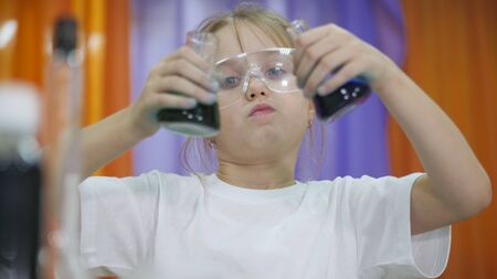 Child is holding flasks of colored liquid. Cute girl in protective transparent glasses on her face....
